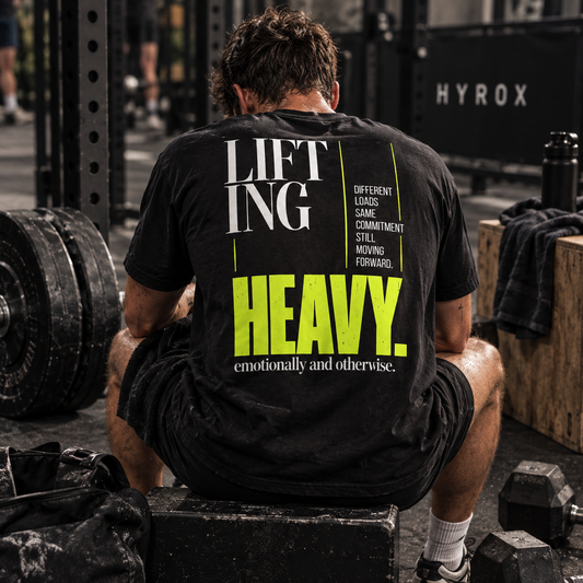Race Scoop Lifting Heavy black graphic tee worn by an athlete recovering between strength training sets, featuring bold back artwork inspired by carrying heavy loads in and out of the gym.